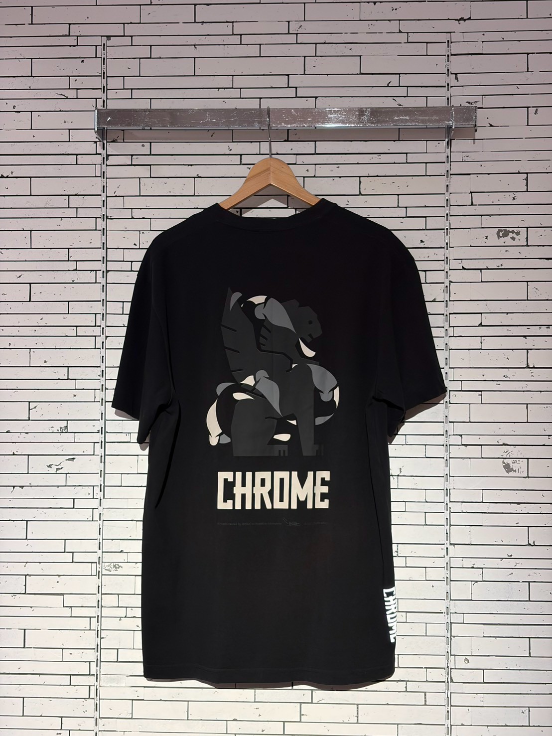 Black T-shirt on a wooden hanger against a white brick wall, displaying a gray abstract graphic and the word CHROME at the bottom center.