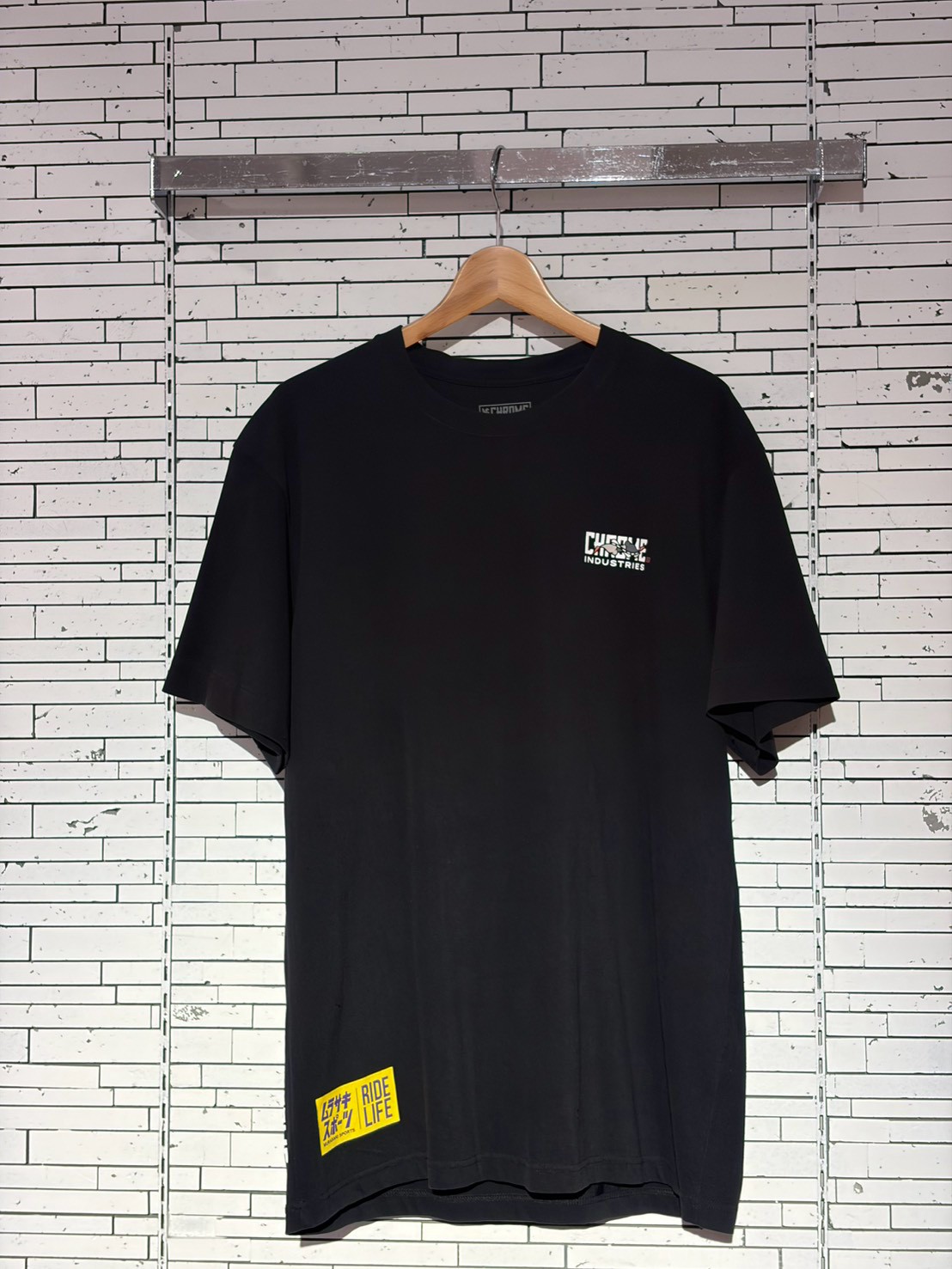 Black short-sleeve T-shirt on a wooden hanger against a white tiled wall; small left-chest graphic logo and a yellow Ride Life patch near the bottom hem.