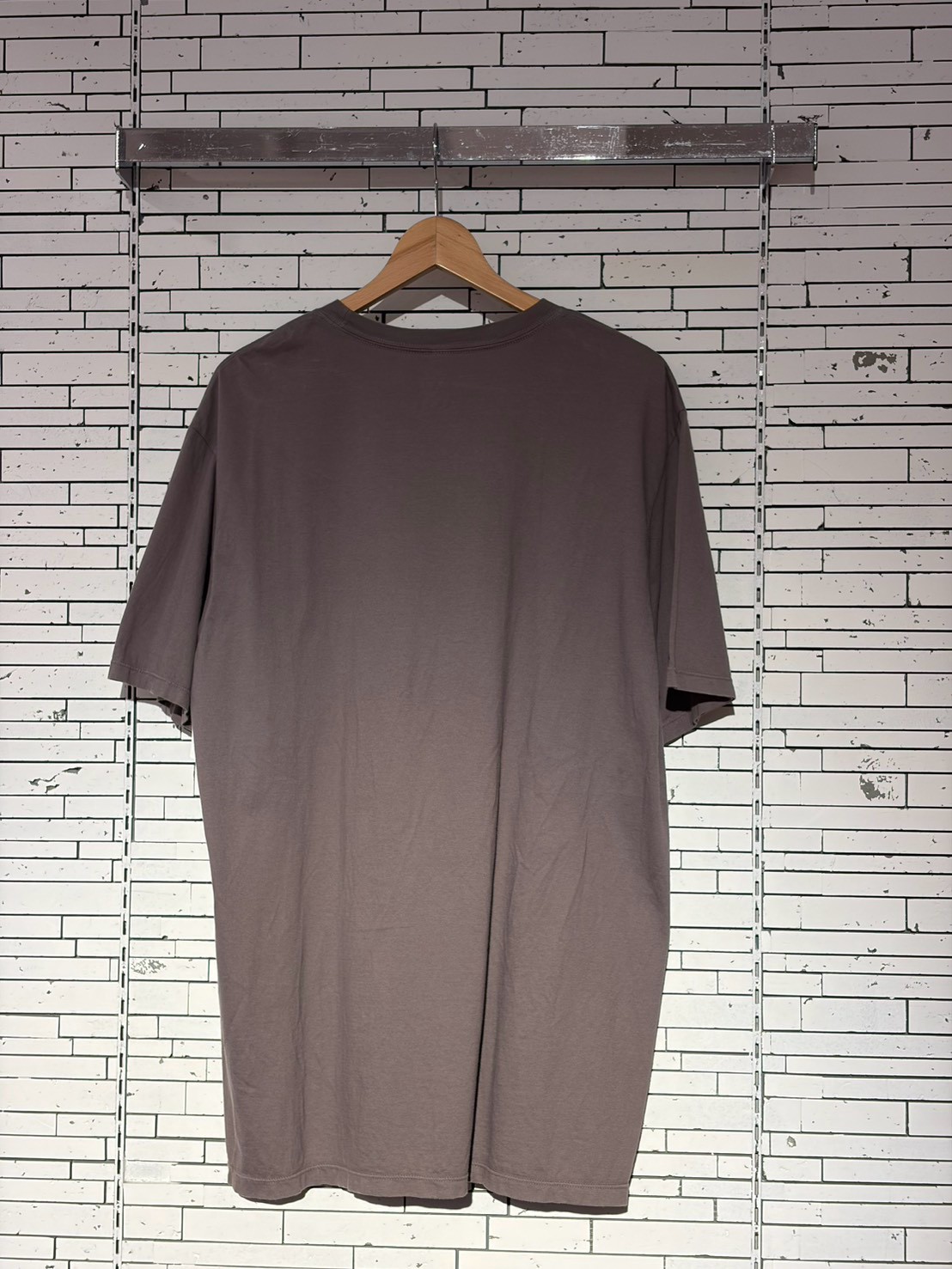 Long, loose taupeT-shirt on a wooden hanger against a white brick wall backdrop.