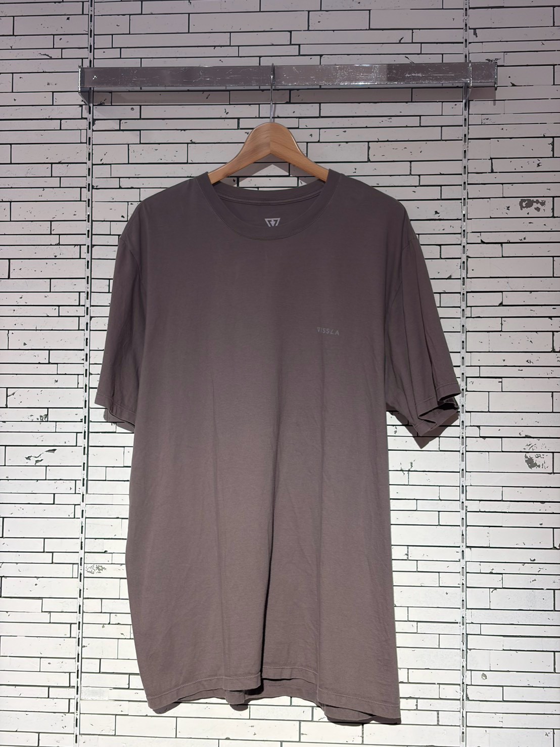 Taupe oversized T-shirt on a wooden hanger against a white-tiled wall, with a small Vissla logo on the chest.