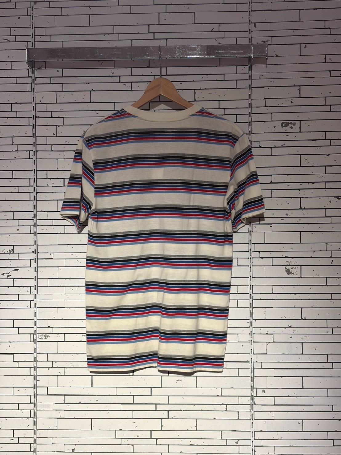 Striped short-sleeve T-shirt on a wooden hanger against a white brick wall. Subtle retail display vibe.