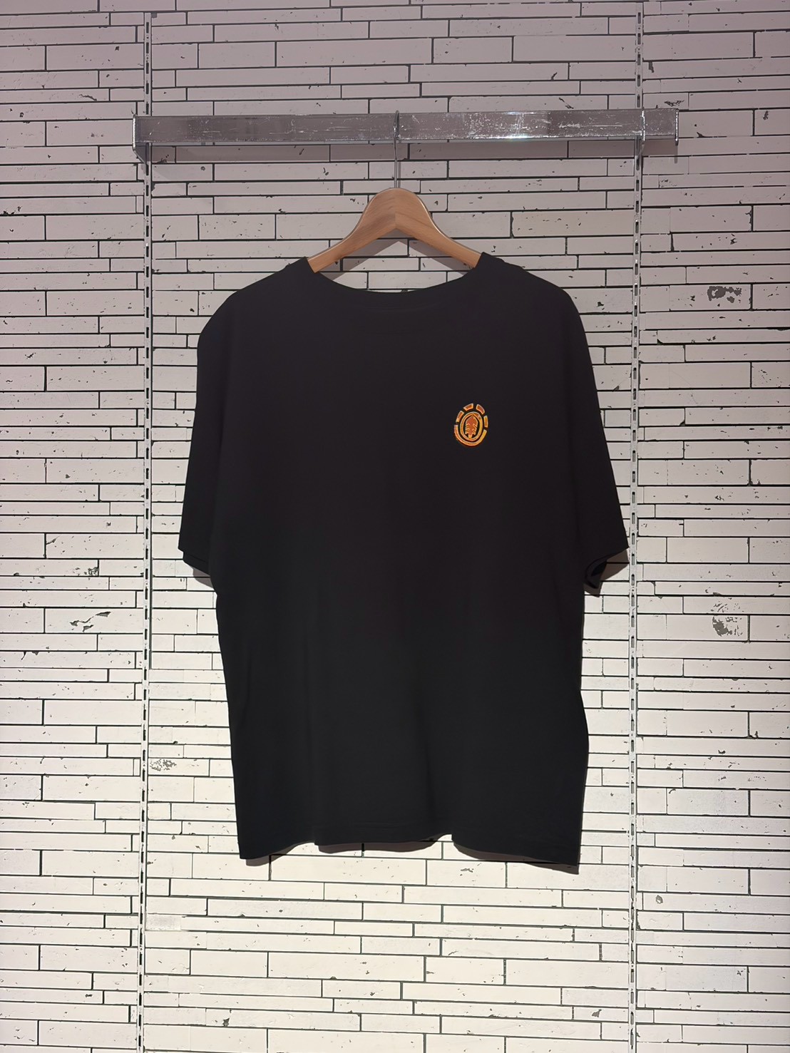 Black t-shirt on a wooden hanger with a small orange circular logo on the left chest, against a white brick wall backdrop.