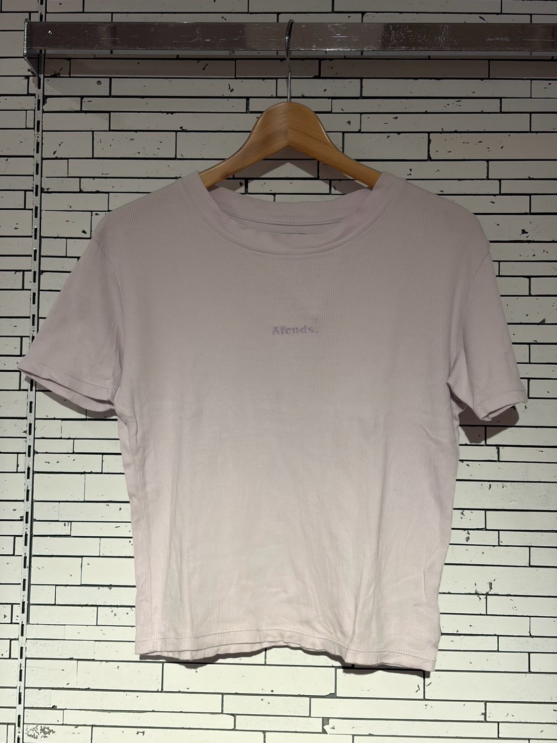 Light pink short-sleeve T-shirt on a wooden hanger against a white brick wall; small chest text/logo visible.