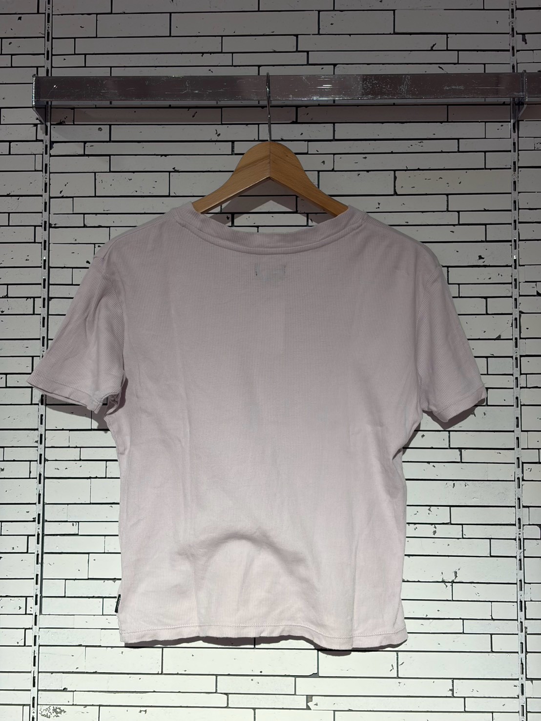 Light pink short-sleeve T-shirt on a wooden hanger against a white brick wall.
