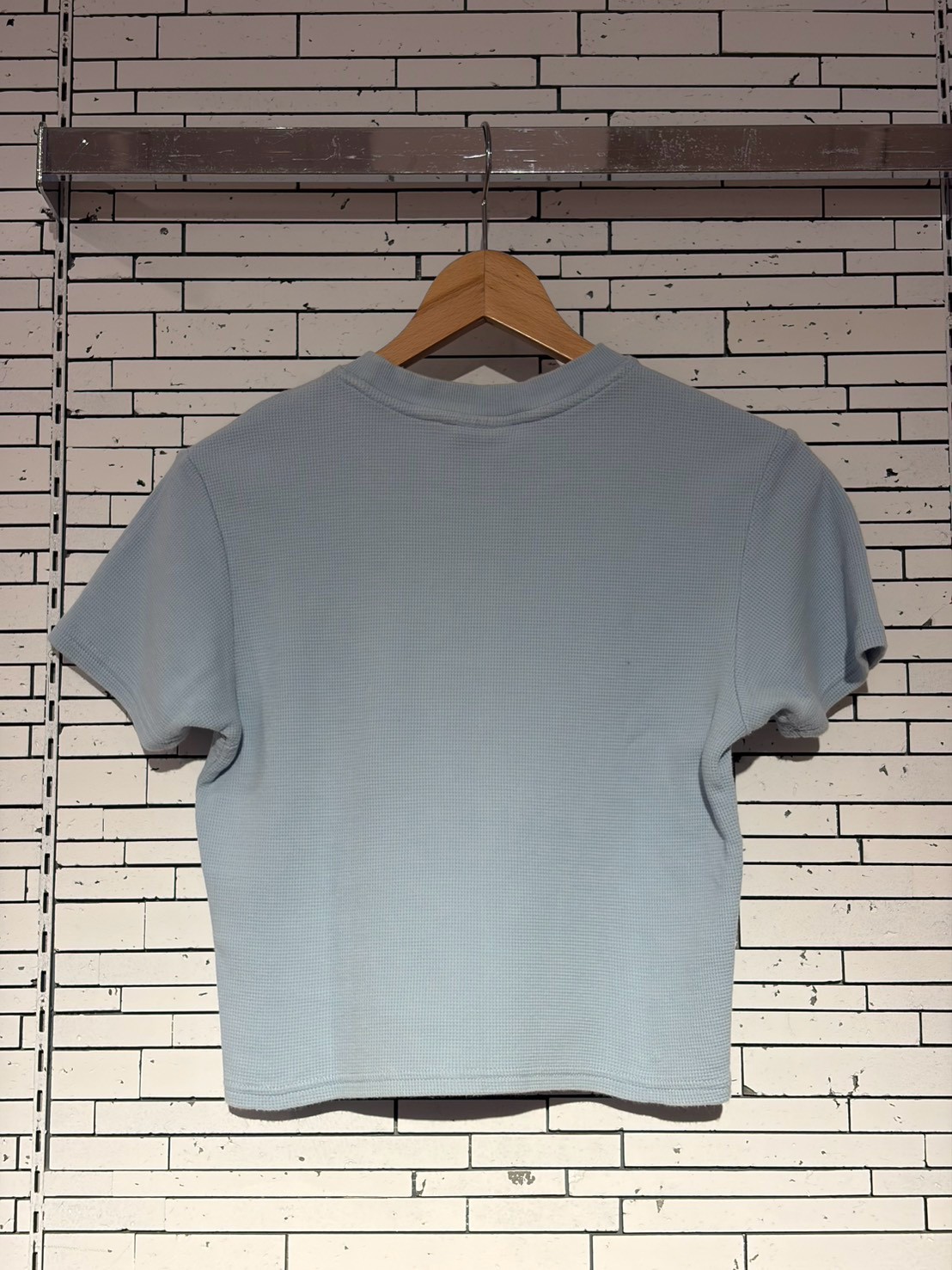Light blue cropped T-shirt on a wooden hanger against a white brick wall.