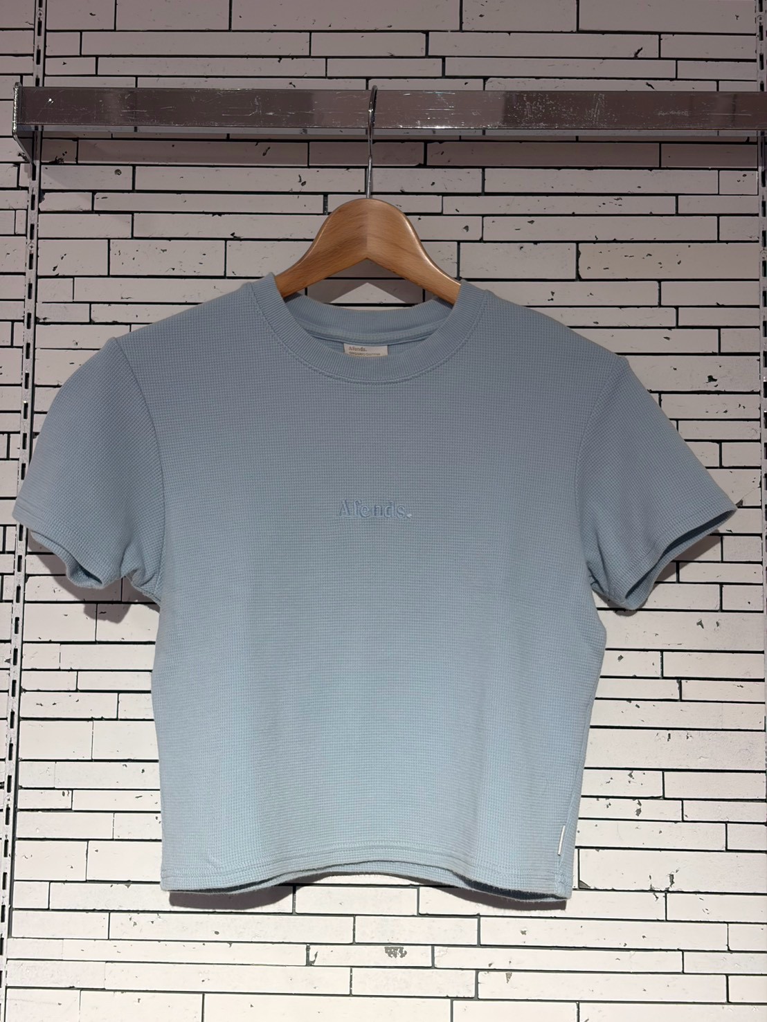 Light blue textured short-sleeve T-shirt on a wooden hanger against a white brick wall.
