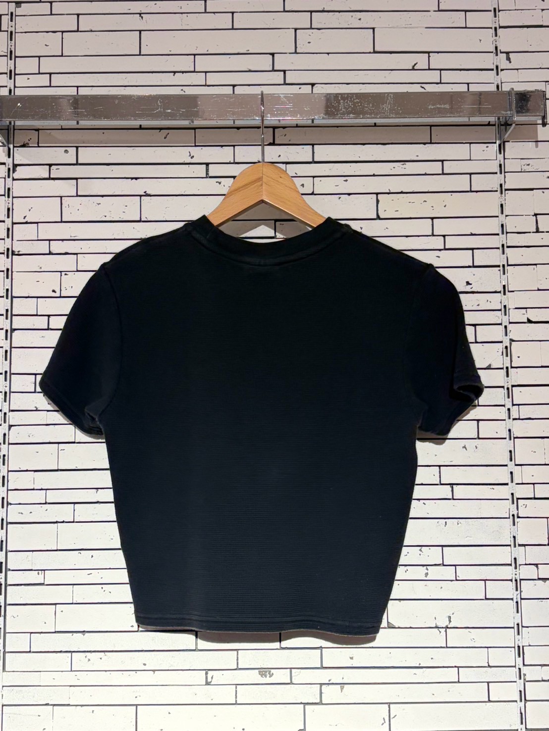 Black short-sleeve crew-neck T-shirt on a wooden hanger against a white brick wall.