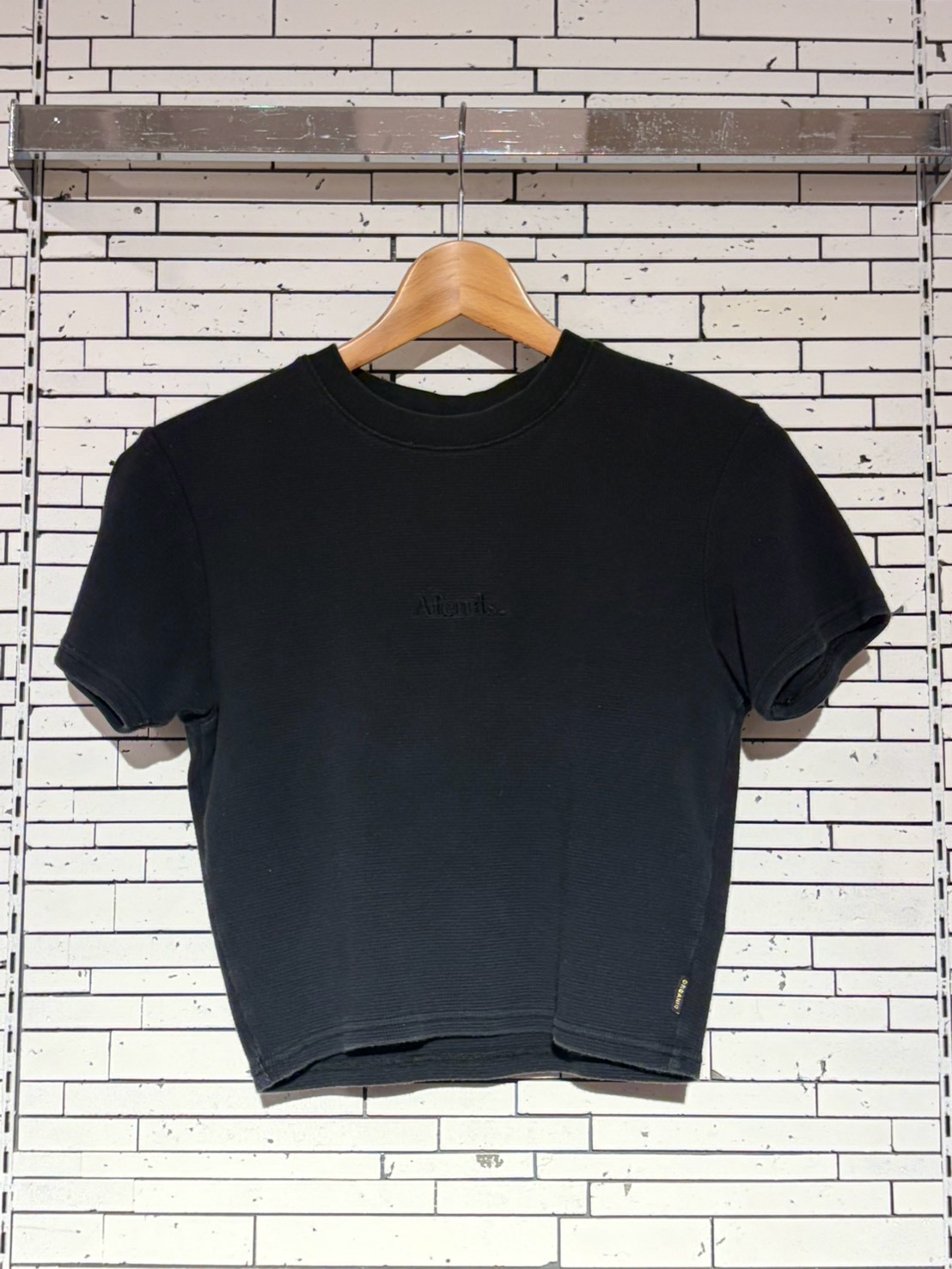 Black short-sleeve crew-neck T-shirt on a wooden hanger against a white brick wall backdrop.