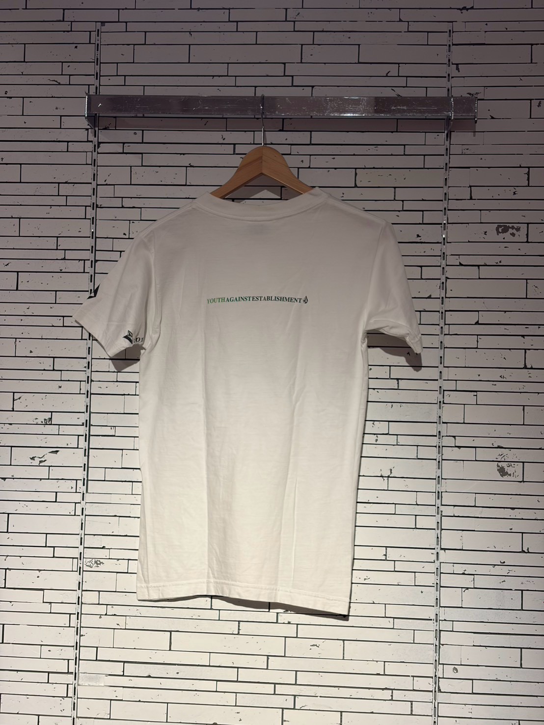 White T-shirt on a wooden hanger against a white brick wall, with a small green slogan reading 'YOUTH AGAINST ESTABLISHMENT' on the chest