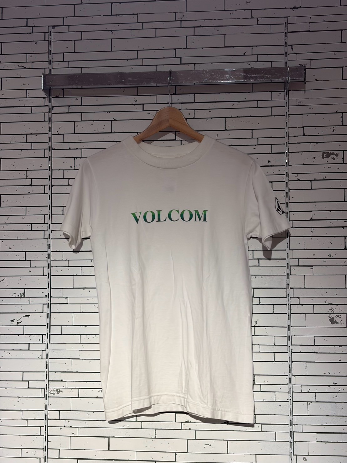 White Volcom T-shirt on a wooden hanger against a white brick wall backdrop.