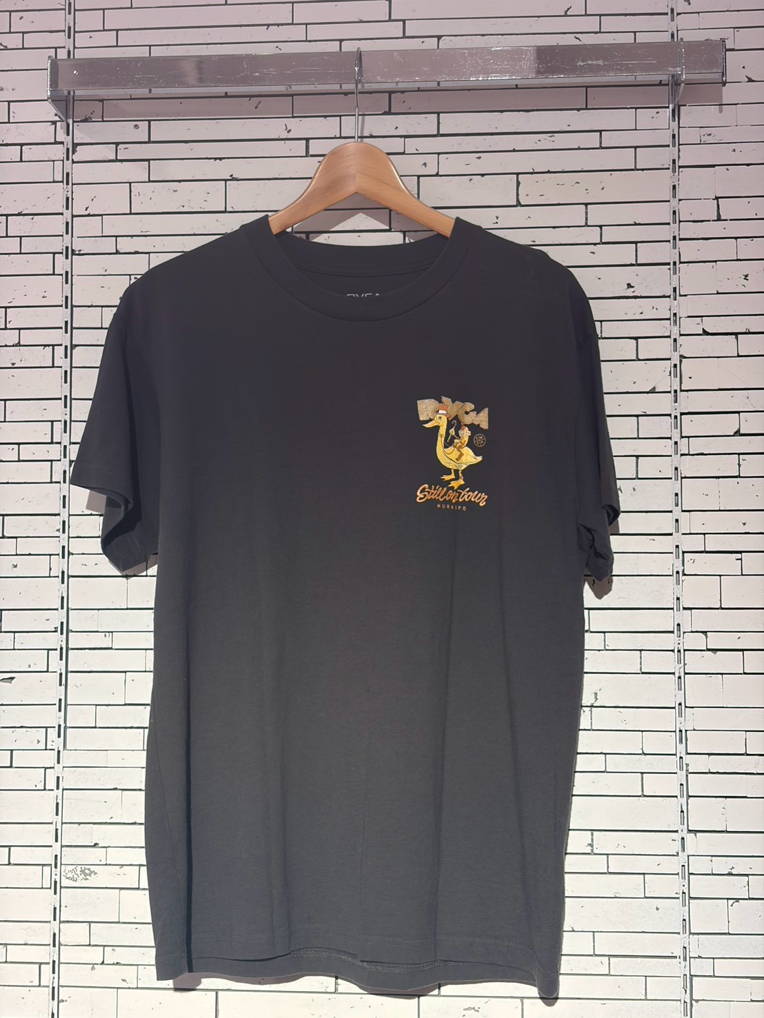 Black T-shirt on a wooden hanger with a small gold rooster graphic and cursive text on the left chest, hanging against a white brick wall.