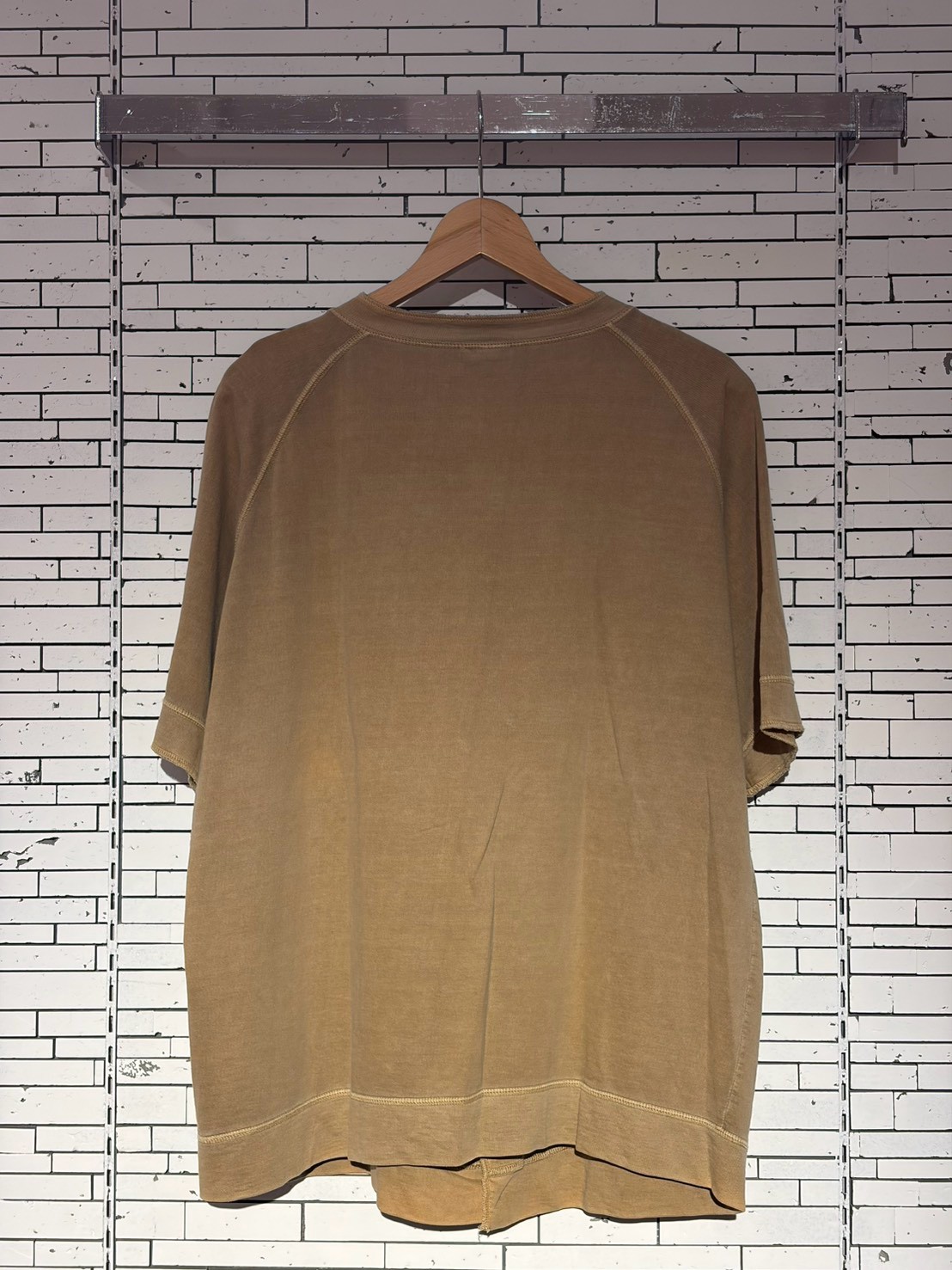 Brown oversized short-sleeve T-shirt on a wooden hanger against a white brick wall, in a retail display.