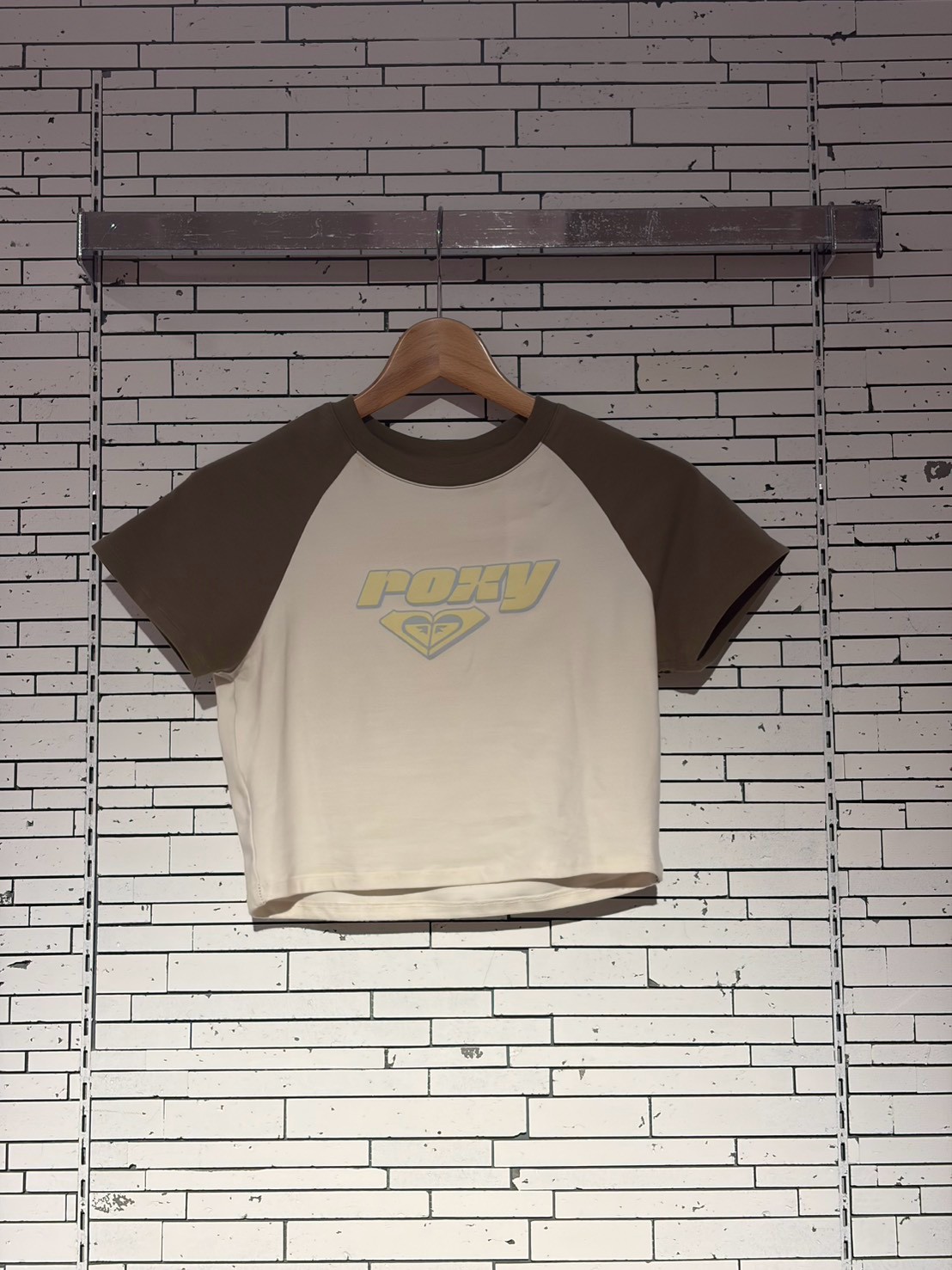 White raglan-sleeve T-shirt on a wooden hanger against a brick wall, with a yellow ROXY logo on the chest.