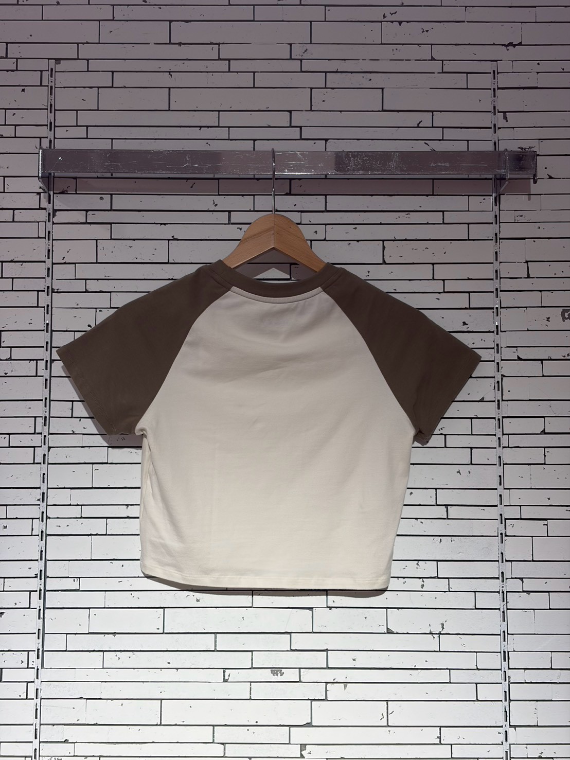 White and brown raglan T-shirt on a wooden hanger against a white brick wall backdrop.