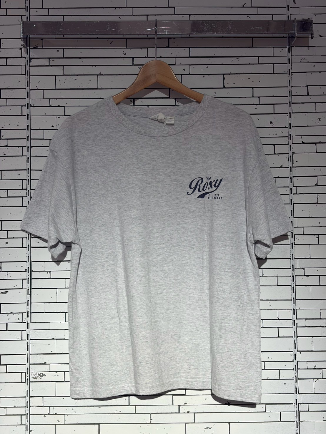 Gray T-shirt on a wooden hanger against a white tiled wall, featuring a small Roxy logo on the left chest.