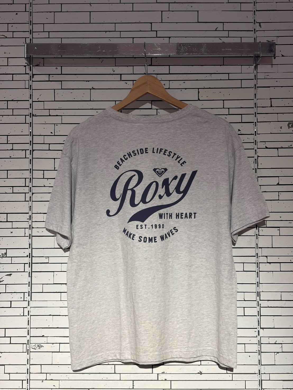 Gray t-shirt on a wooden hanger with a large Roxy logo and 'Beachside Lifestyle' text, hanging on a white brick wall background.