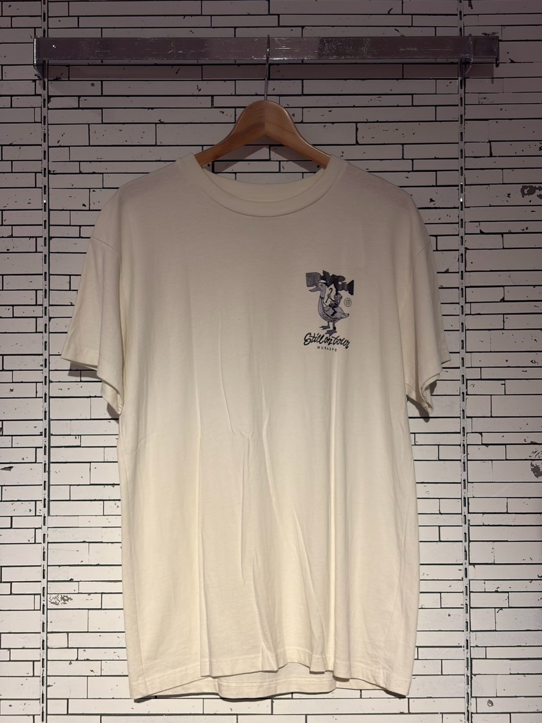 White T-shirt on a wooden hanger against a white brick wall, with a small cartoon graphic on the left chest area.