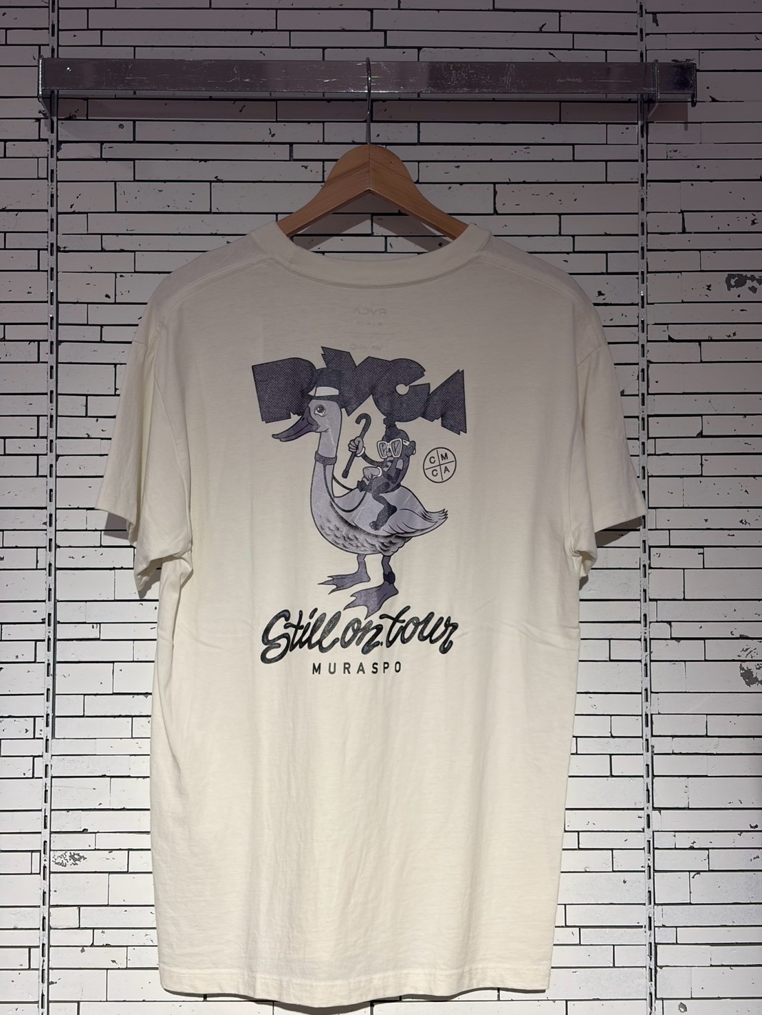Cream T-shirt on a hanger against a white brick wall, featuring a cartoon duck and a rider with the caption 'Still on tour' and 'MURASPO'.