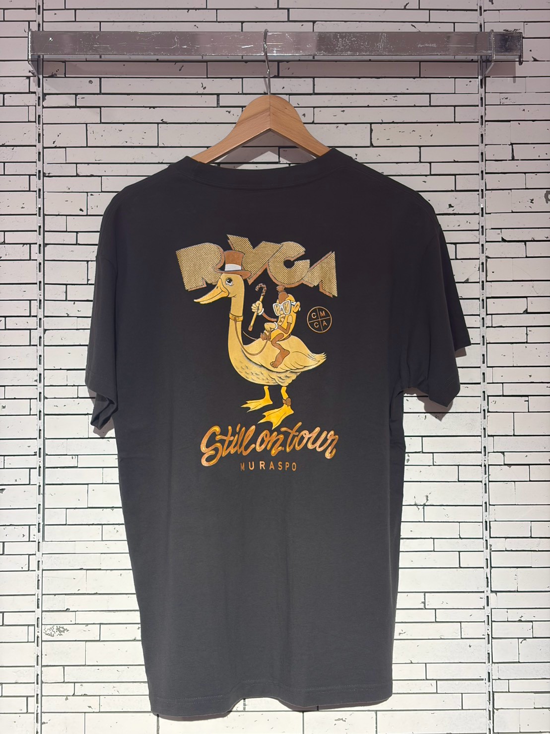 Black T-shirt on a hanger against a white brick wall, featuring a vintage cartoon print of a duck with a rider and the text 'Still on tour'.