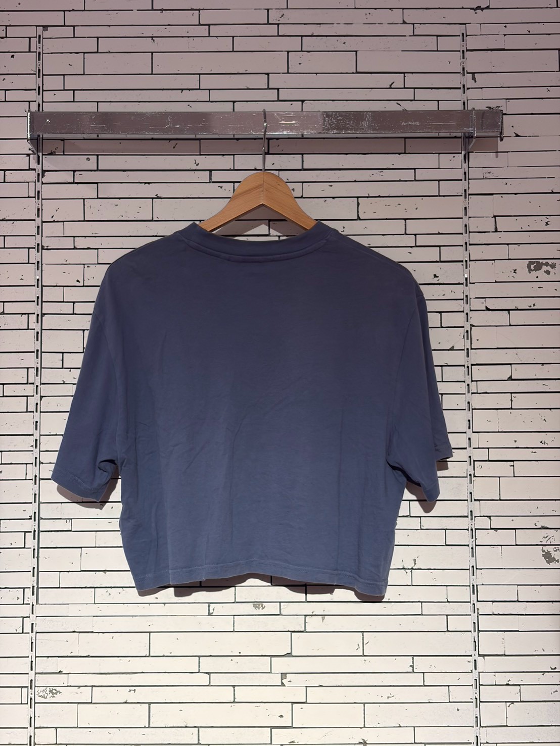Blue short-sleeve T-shirt on a wooden hanger against a white brick wall, displayed on a metal rack.
