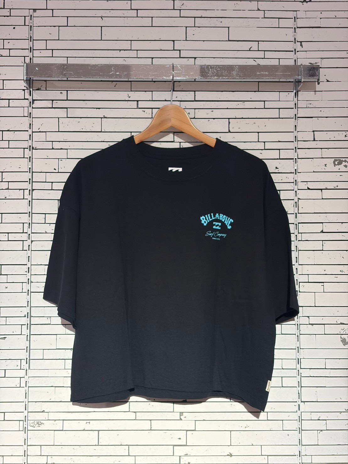 Black oversized T-shirt on a wooden hanger against a white brick wall, featuring a small blue Billabong logo on the left chest area for branding.