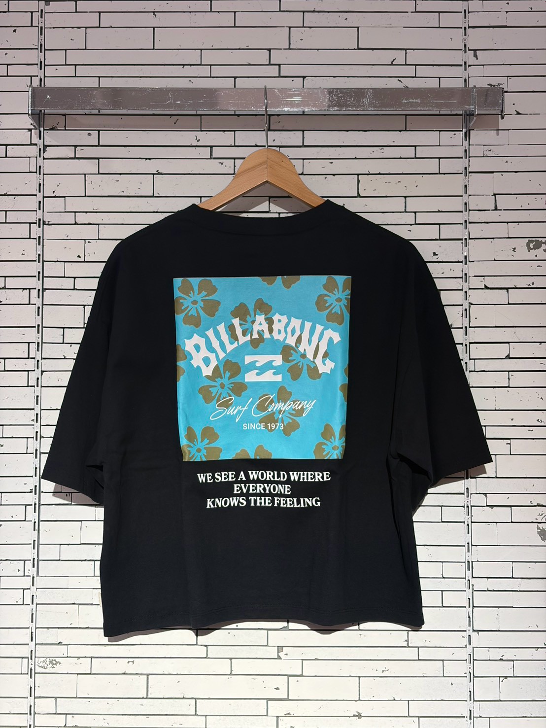 Black T-shirt hanging on a wooden hanger with a large turquoise graphic back panel featuring hibiscus flowers and the words 'Billabong Surf Company' and 'Since 1973' with a film-style script; slogan below reads 'We see a world where everyone knows the feeling'.