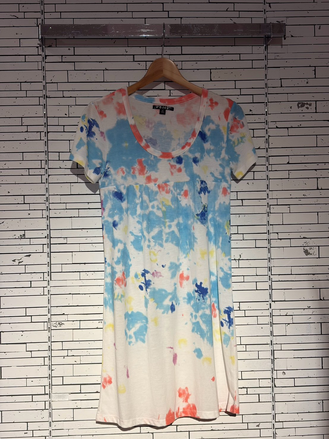 Tie-dye short-sleeve dress on a wooden hanger against a light, striped wall, featuring blue, orange, and yellow splashes.