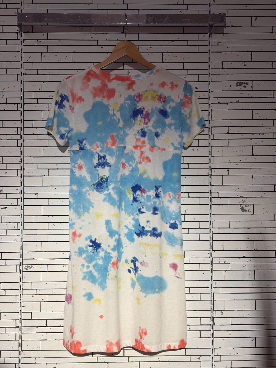 Tie-dye dress on a wooden hanger against a white brick wall; colorful blues, reds, and yellows form abstract patterns.
