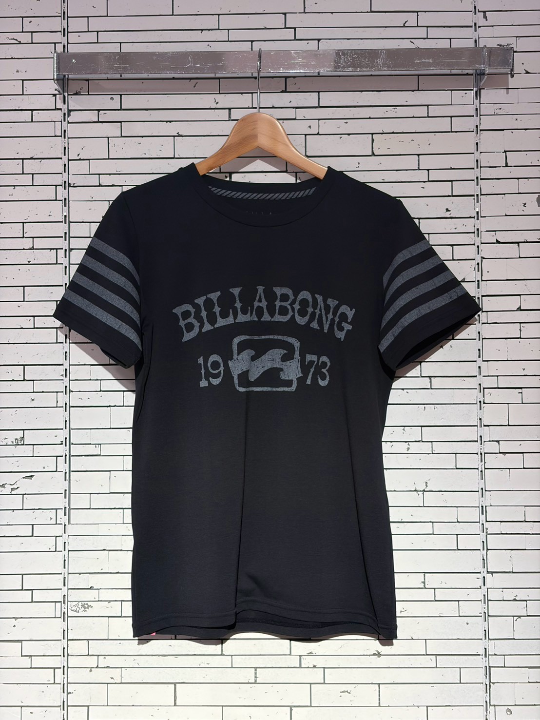 Black short-sleeve Billabong T-shirt on a wooden hanger against a white brick wall, featuring a faded logo and striped sleeve cuffs.