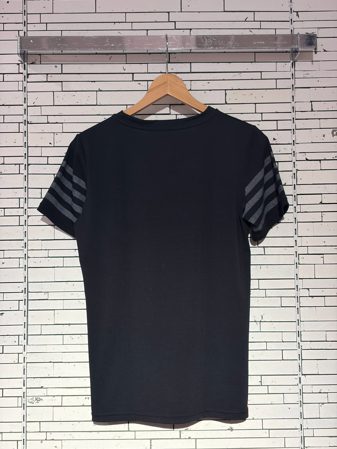Black short-sleeve T-shirt on a wooden hanger against a white brick wall; sleeves have gray stripes