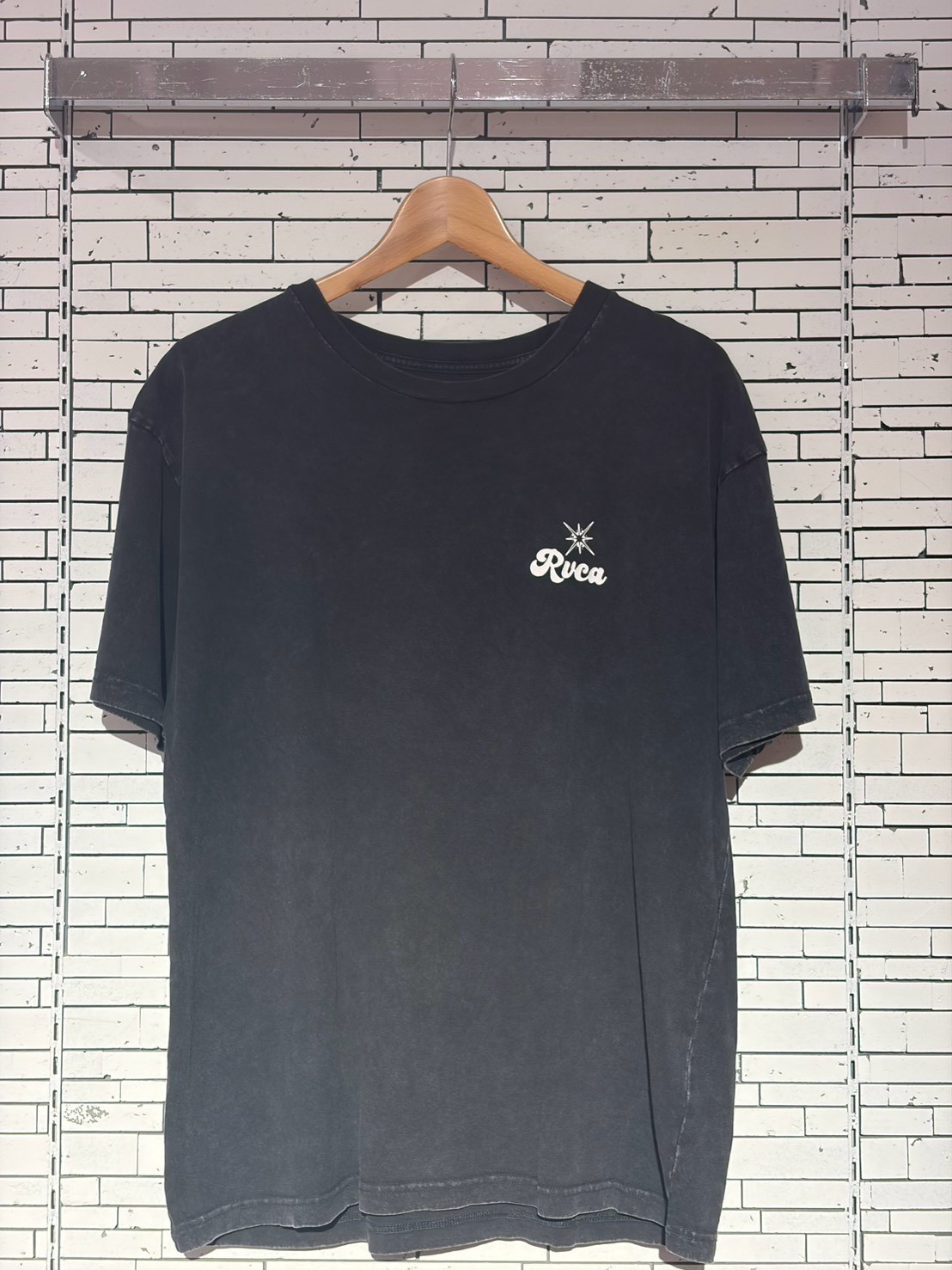 Dark washed short-sleeve T-shirt on a wooden hanger against a white tile wall, small RVCA logo with a star on the left chest area.
