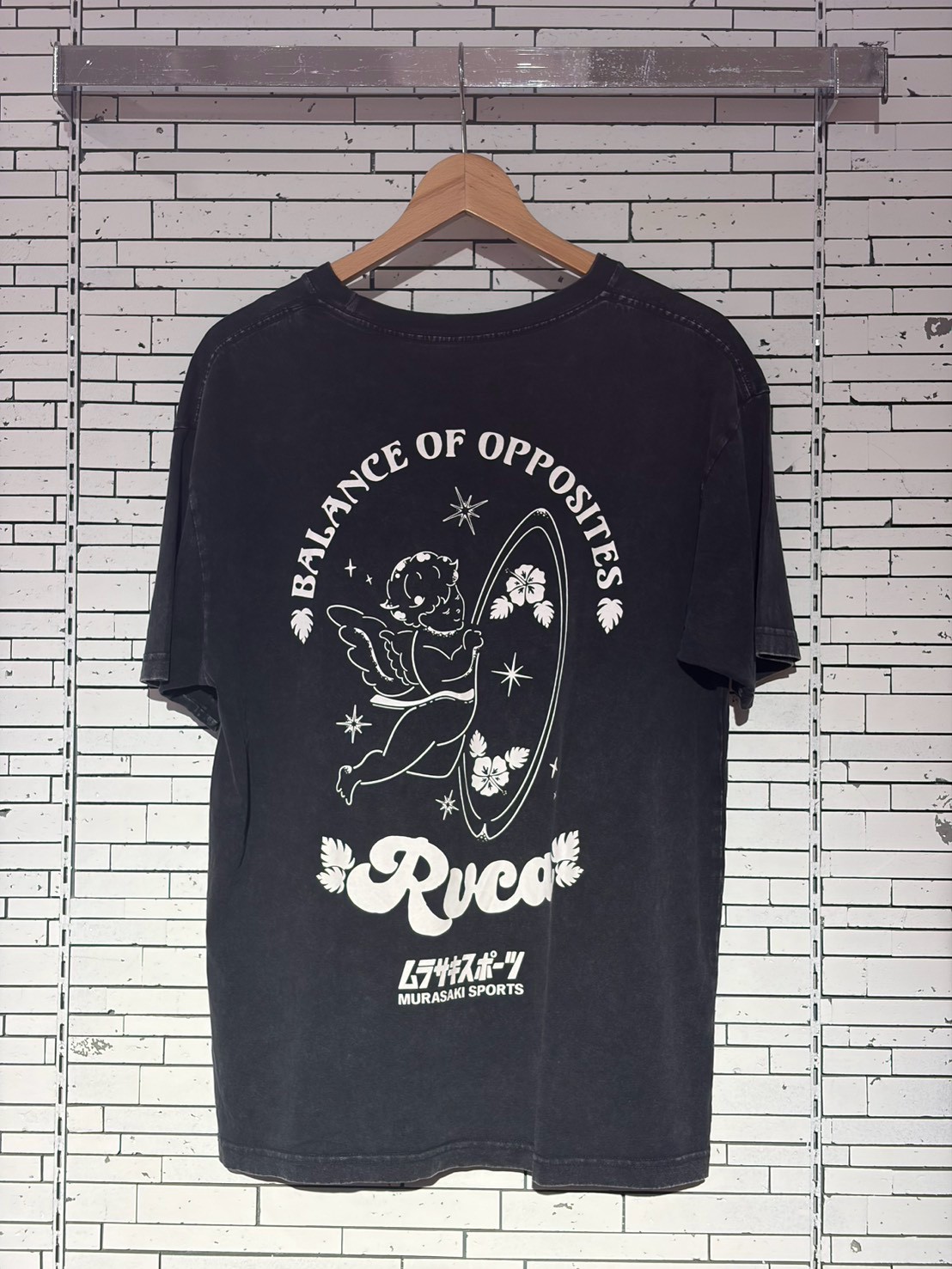 Black T-shirt on a wooden hanger against a white brick wall; front shows a white retro graphic with a cherub by a surfboard, hibiscus flowers, and the words 'Balance of Opposites' and 'RVCA' with 'Murasaki Sports' text at the bottom.