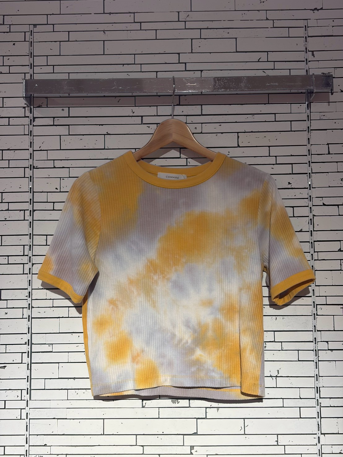 Tie-dye yellow and white short-sleeve T-shirt on a wooden hanger against a white brick wall.