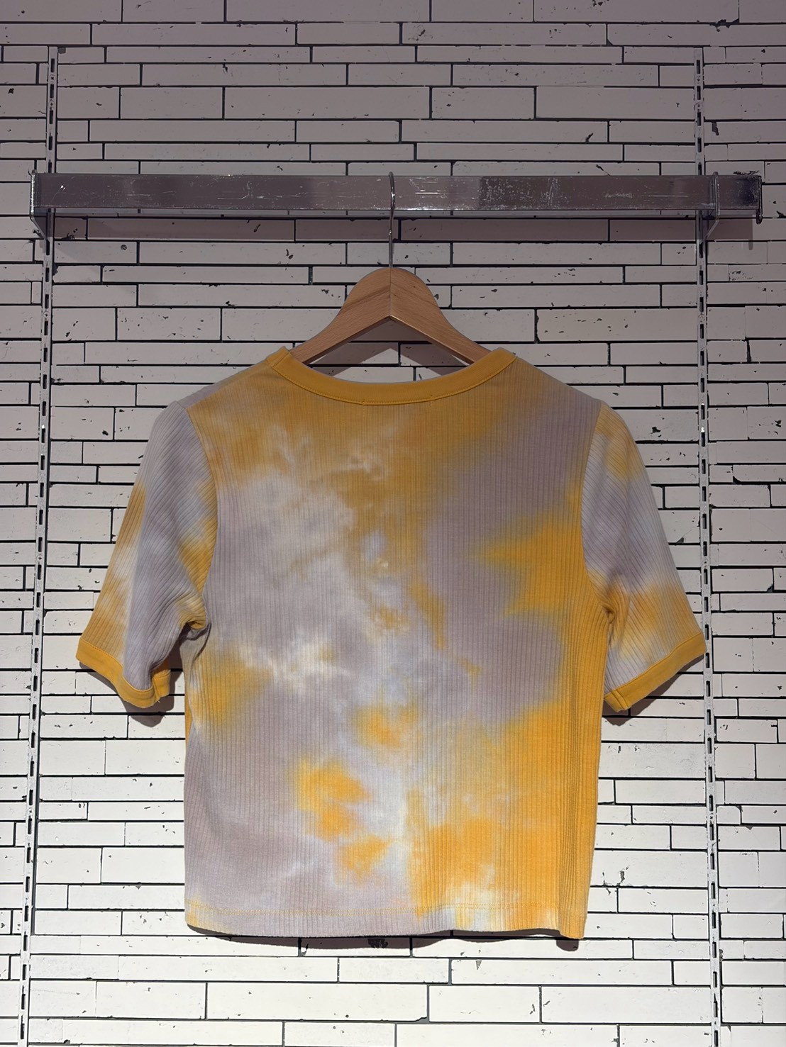 Yellow and gray tie-dye short-sleeve T-shirt on a wooden hanger against a white brick wall, full view.