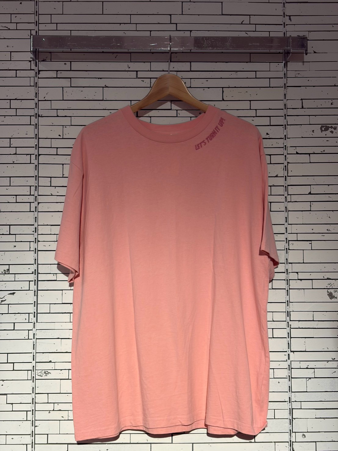 Pink oversized T-shirt on a wooden hanger against a white brick wall, with small text near the shoulder saying 'LET'S TURN IT UP'.