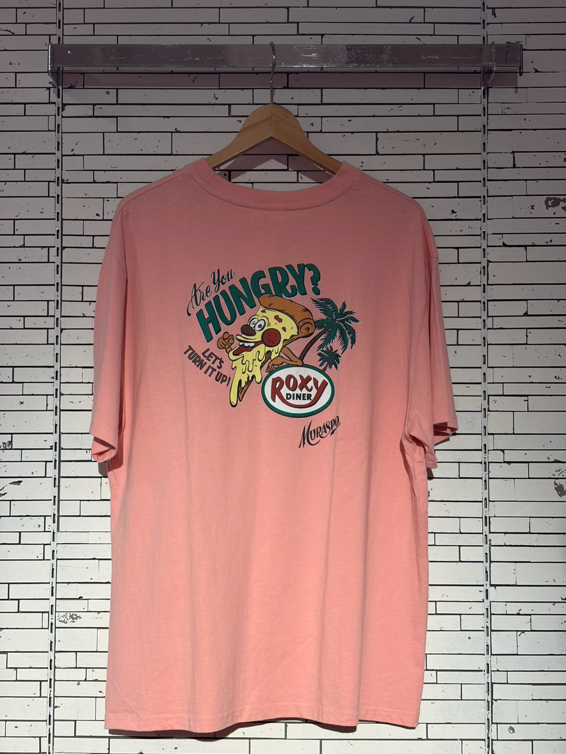 Pink T-shirt on a hanger with a cartoon pizza slice and palm tree graphic reading 'Are you hungry? Roxy Diner'