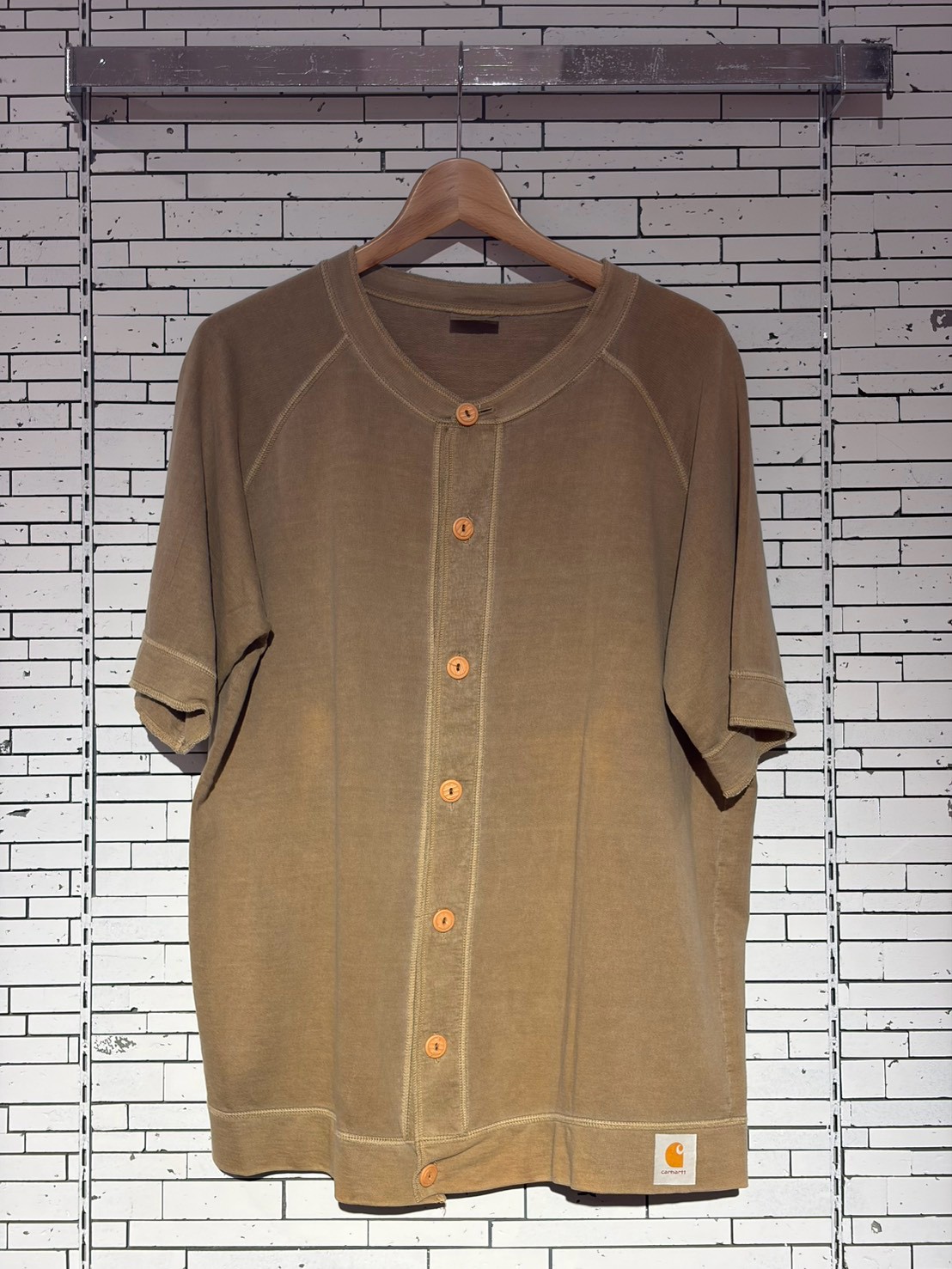 Brown short-sleeve button-front shirt on a wooden hanger against a white brick wall; Carhartt logo tag visible at the bottom hem