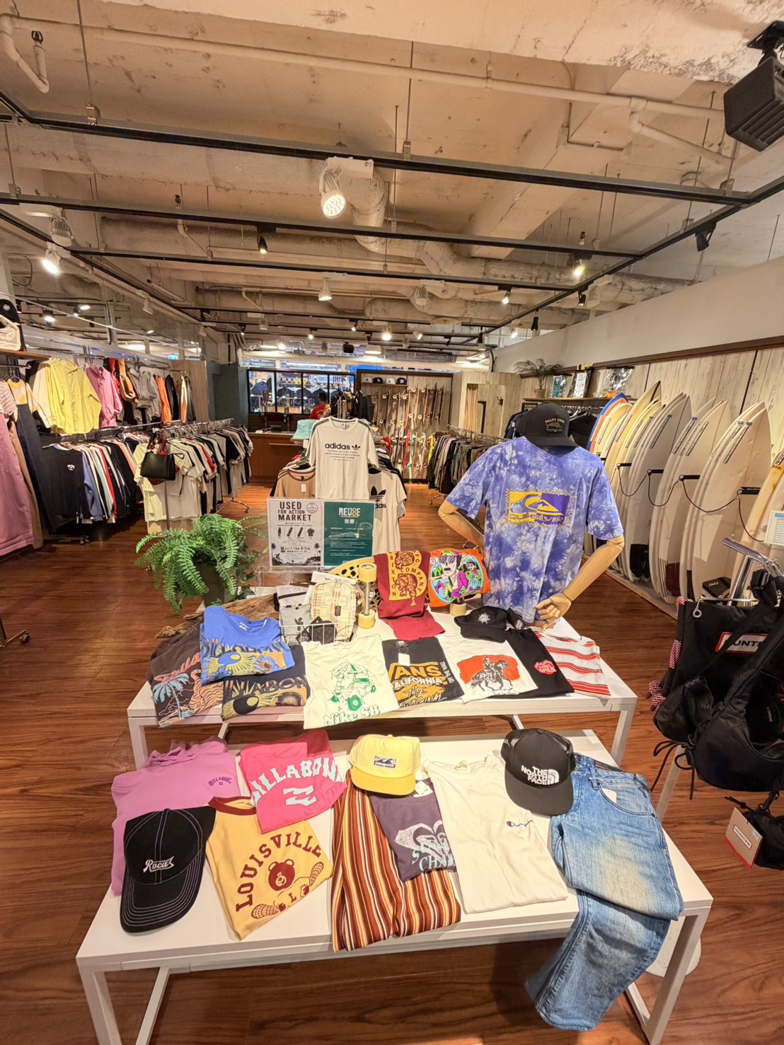 Inside a sunny casual clothing shop with racks of jackets and shirts, a central table displays graphic tees and hats, plus a blue tie-dye shirt on a mannequin; surfboards line the wall to the right.